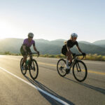 Two cyclists riding on an open road at sunrise with mountains in the background.