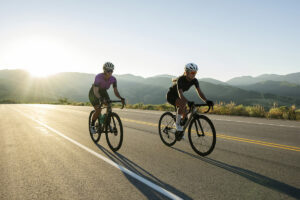 Two cyclists riding on an open road at sunrise with mountains in the background.