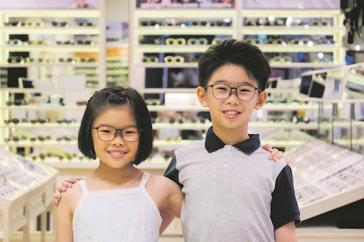 Two_children_wearing_prescription_glasses_smiling_in_an_optometry_clinic_with _a_wide_range_of_eyewear_displayed_in_the_background.